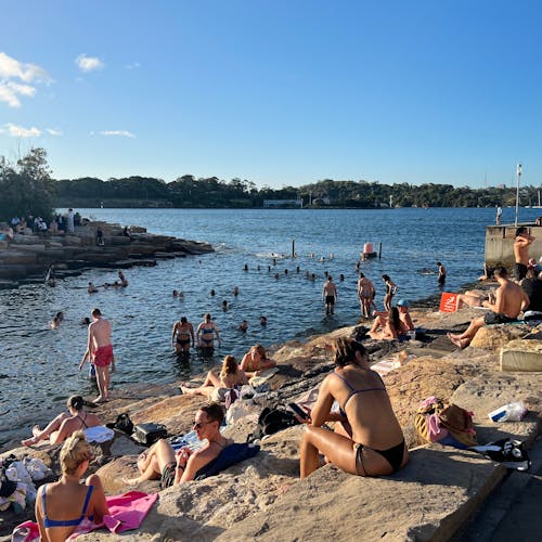 A photograph of bathers at a Sydney Harbour swimming spot. People swim, as well as sit on sandstone terraces. The sun is shining and a blue sky with small clouds is visible above.