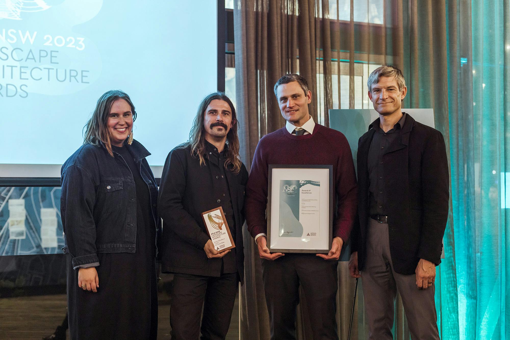 A photograph of four people on an awards stage, holding a small square trophy and a framed certificate. 