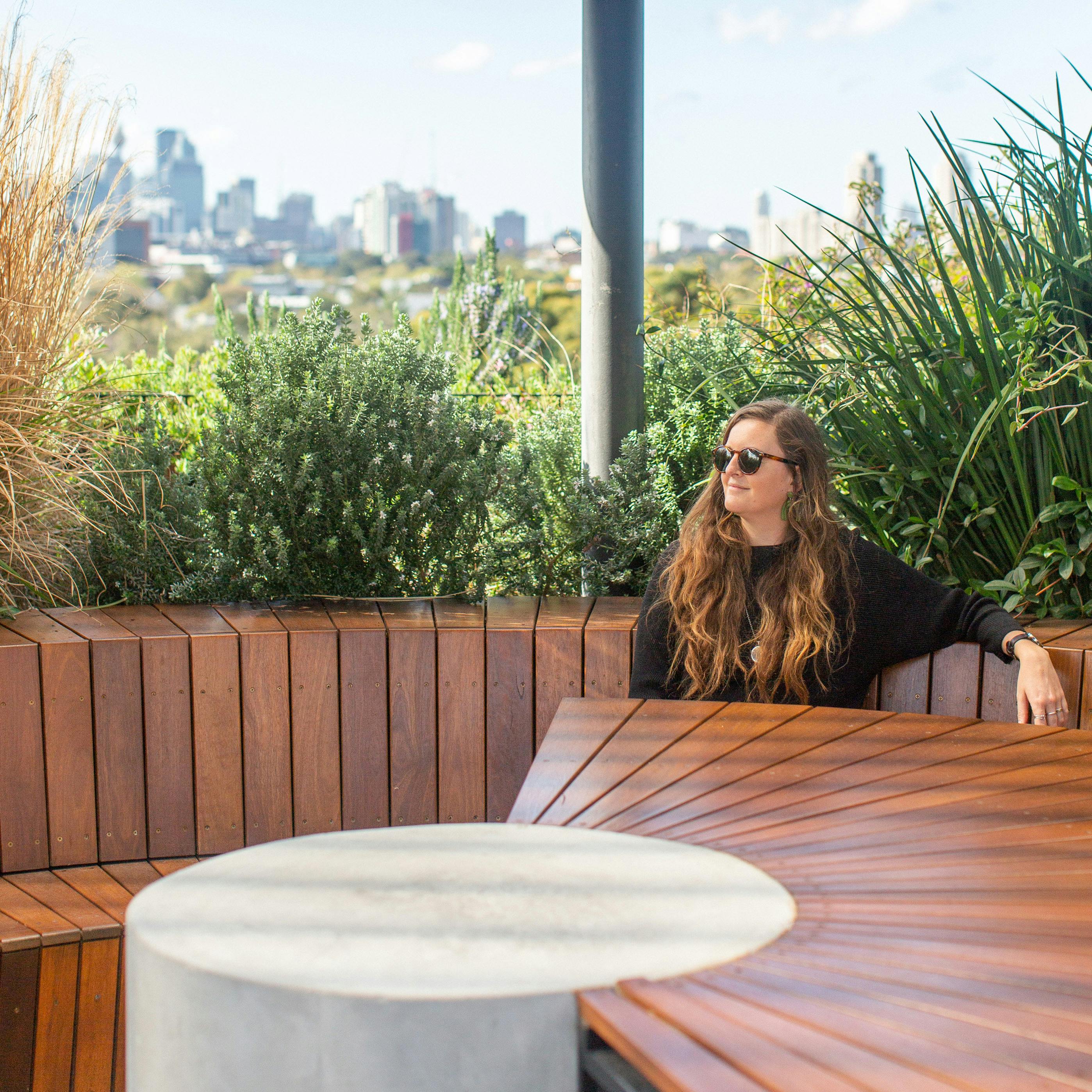 A photograph of a person relaxing in a rooftop garden. They are sitting on a curved seat, at a curved table, and wearing sunglasses. Lush green planting and a glimpse of Sydney's skyline make up the background.