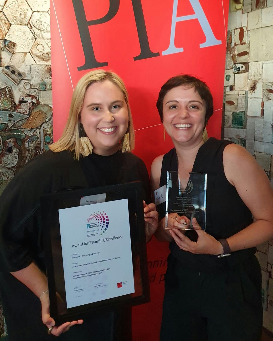 A photograph of two smiling people, one is holding a framed certificate, the other is holding a clear trophy. 