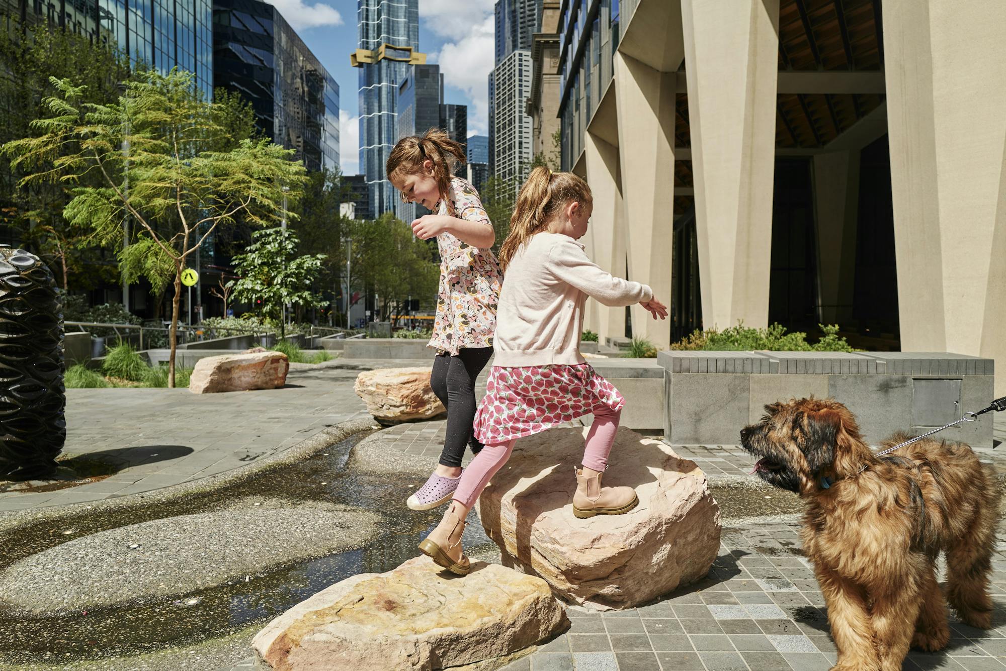 A photograph of two children playing on sandstone rocks in an urban park. A landscaped water channel and banksia sculpture can be seen on the left, and there is a dog on the right. In the background there are trees, plants, and glimpses of a city skyline.