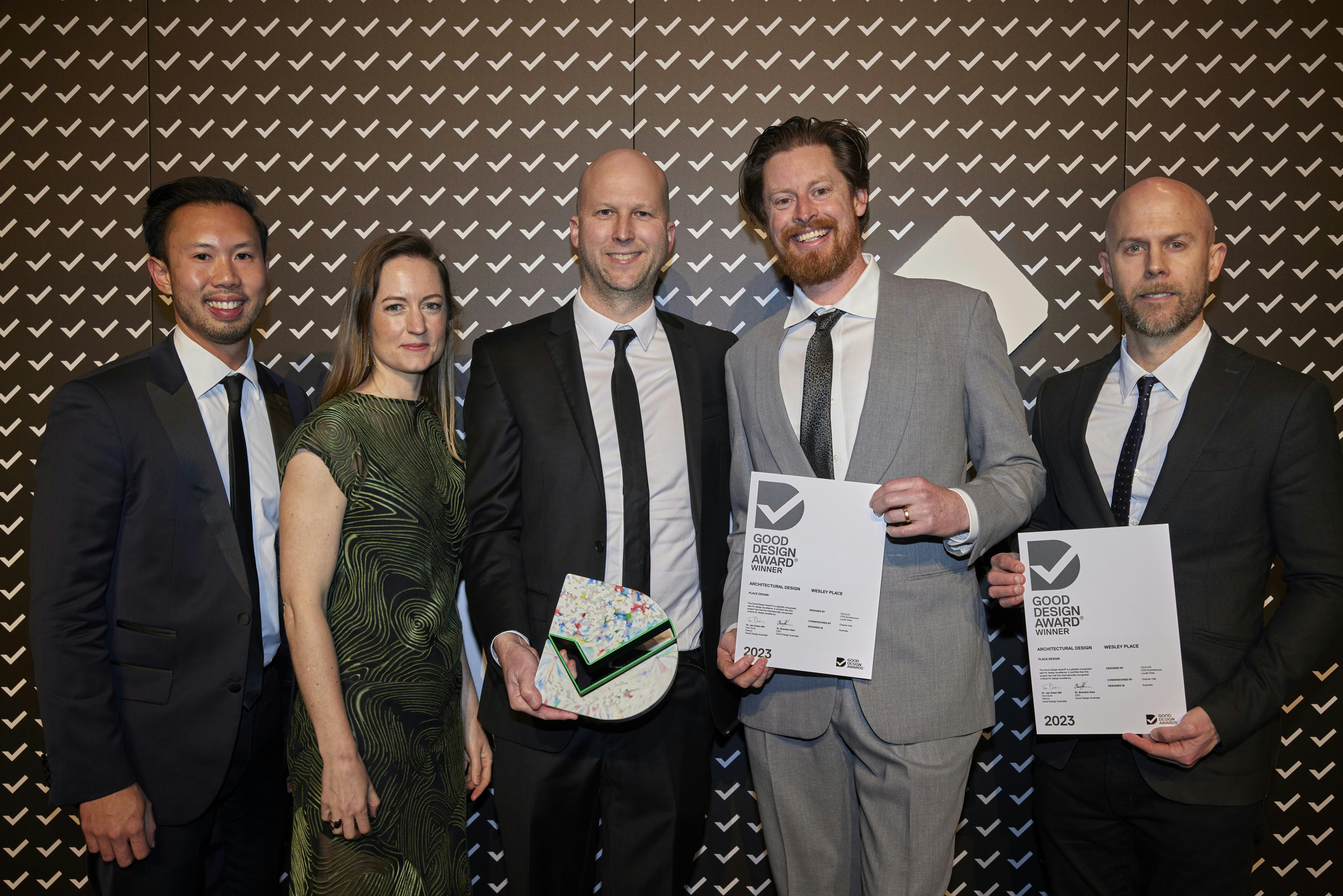 A photograph of five people in front of a media wall. One is holding a trophy, two are holding award certificates.