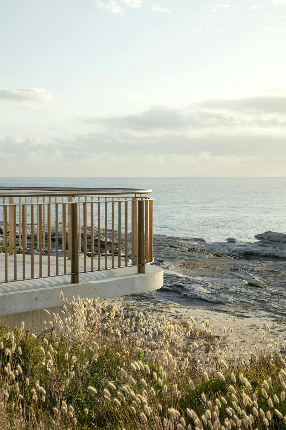 A photograph of the end of curved viewing platform, surrounded by native grasses and rocky cliffs, with a pastel sea and soft cloudy sky in the background.
