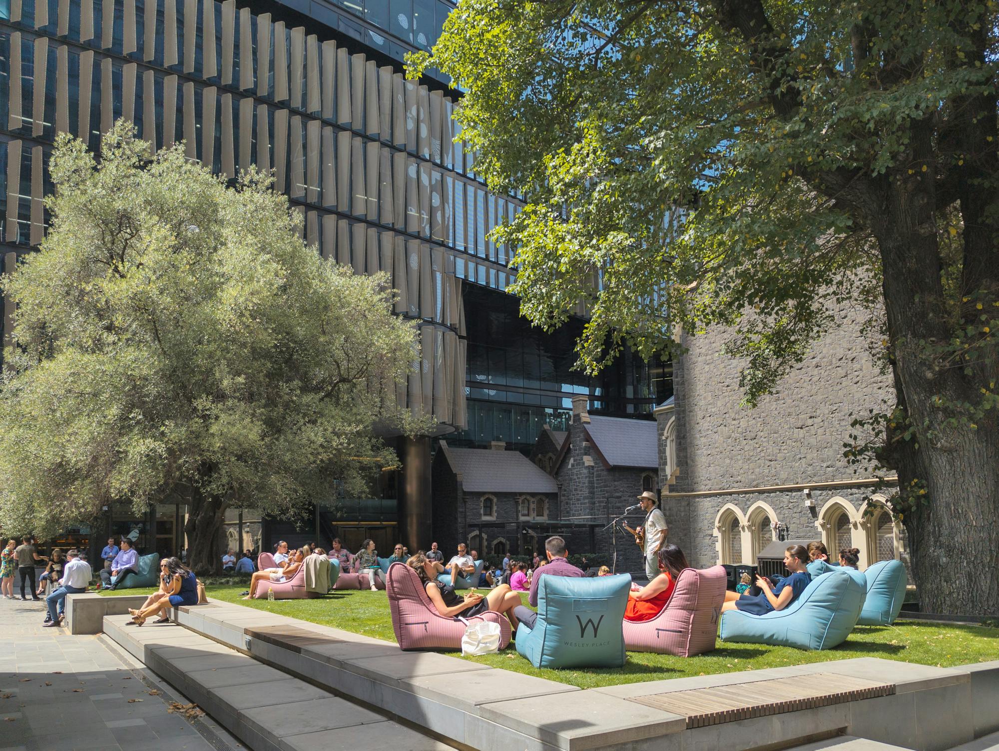 A photograph of an urban green square, there are two large trees, and a crowd of people sitting on pink and blue bean bag chairs listening to a live musician.