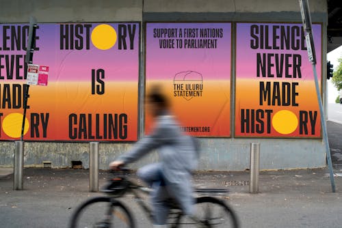 A digital mock-up of Uluru Statement Posters on an urban wall reading "History is Calling" and "Silence Never Made History" with a blurry cyclist passing in the foreground.