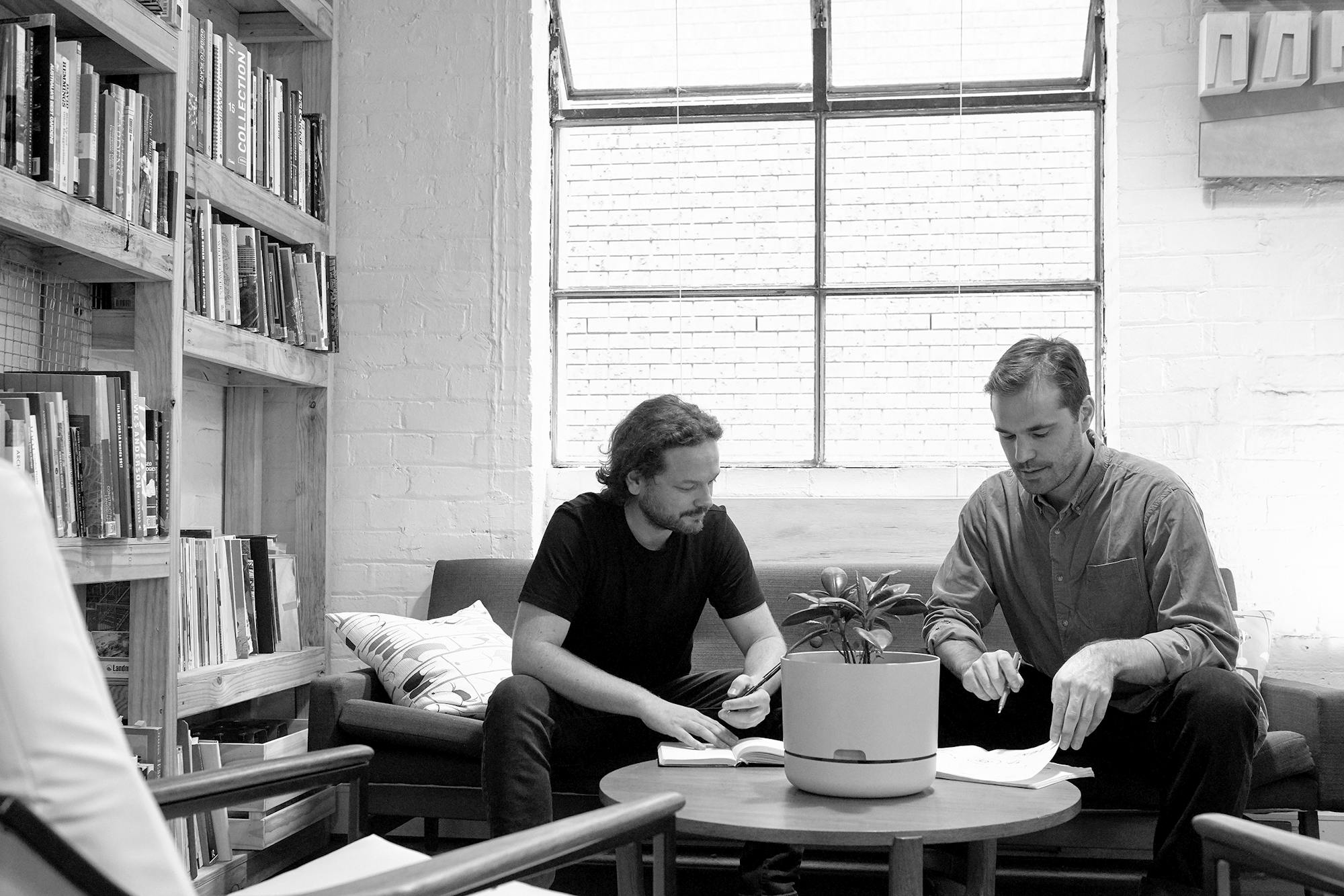 A black and white photograph of two landscape architects sitting on a couch in a warehouse studio. There is a large window behind them, a bookshelf to the left, and a coffee table with a pot plant in the foregound.