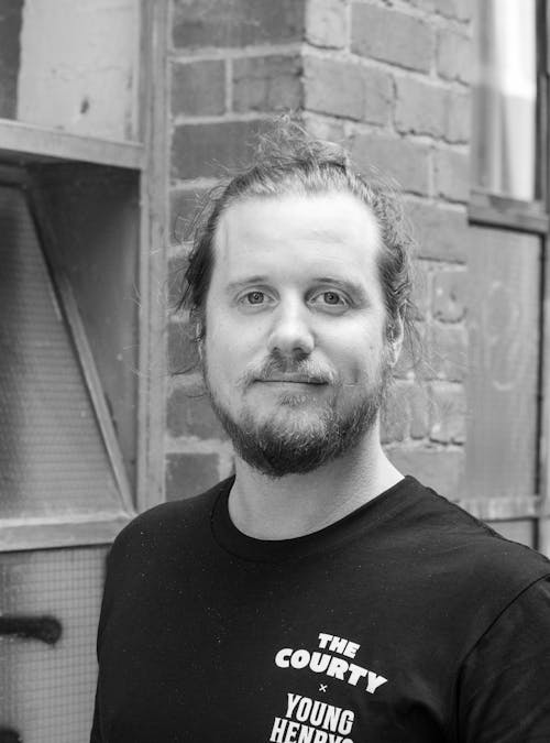 A black and white headshot of a person in-front of a brick wall, wearing a t-shirt that says 'The Courty'