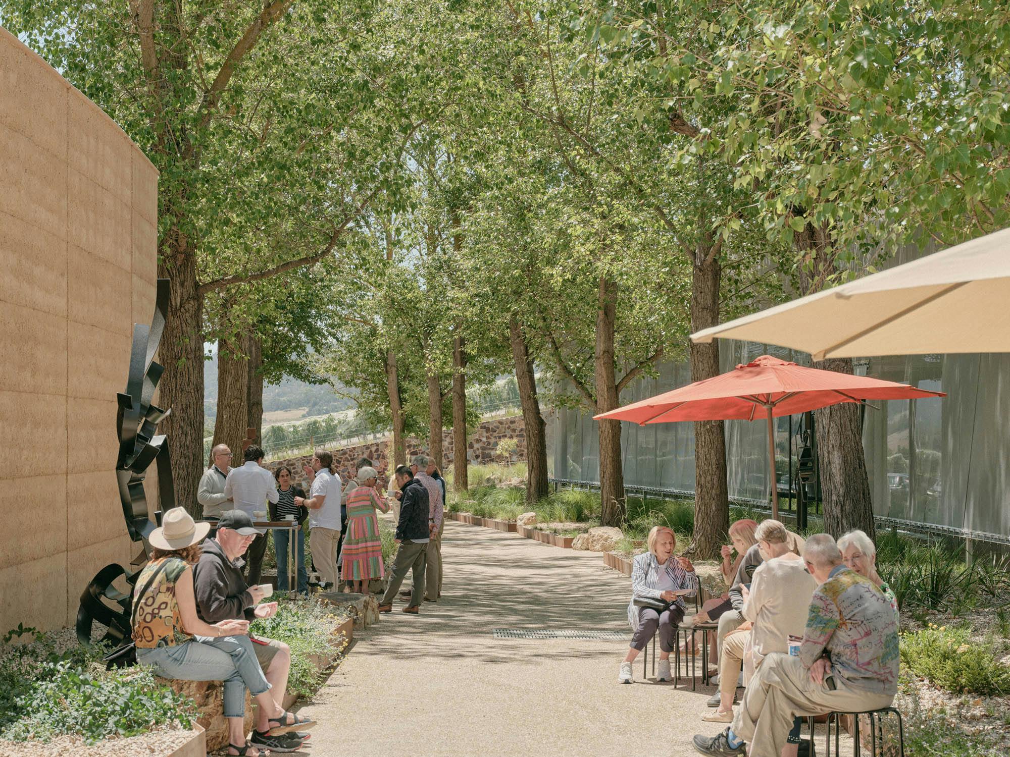 A photo of people socialising in a laneway-like space between two buildings under the shade of trees and umbrellas.