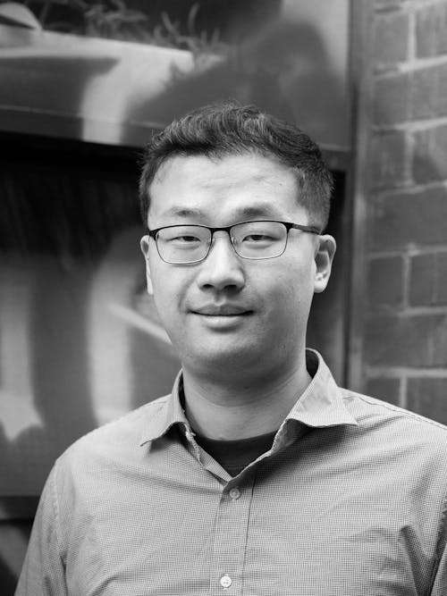 A black and white headshot of a person wearing glasses. The background is an gritty, urban wall.