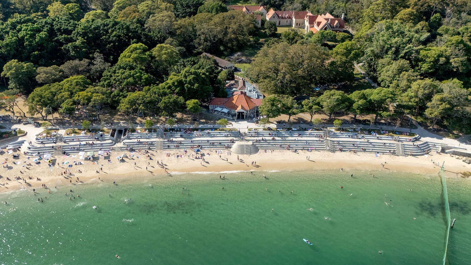 A drone photograph looking down on a sunny beach with a tiered seawall. There are heritage buildings near the water.