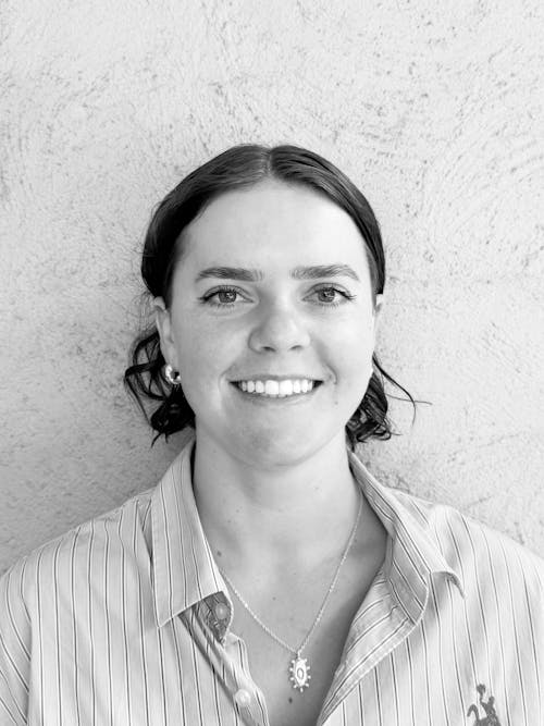 A black and white headshot of a person smiling at the camera. They are wearing a button up shirt, earrings, and a silver organic shaped pendant.