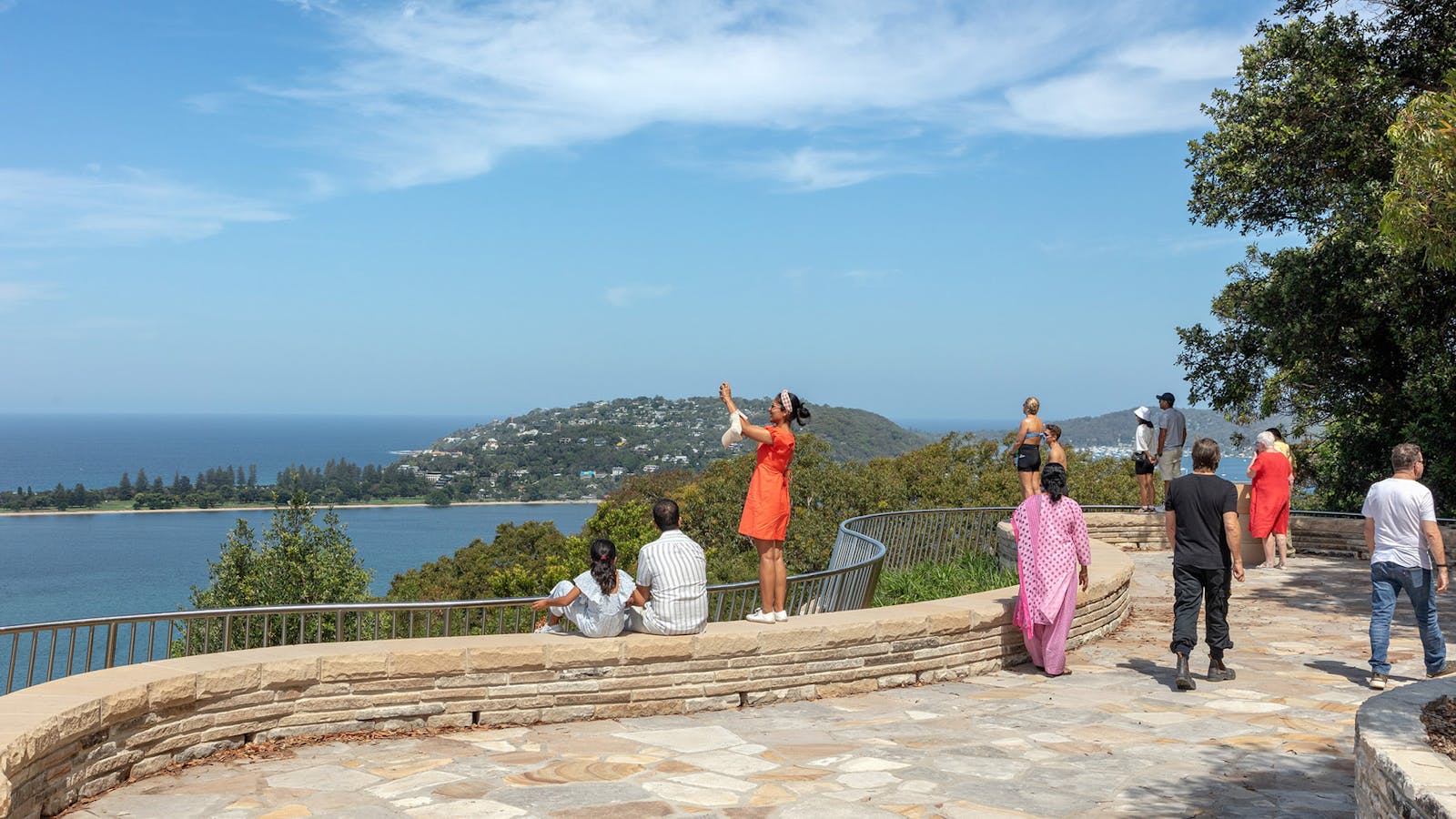 A photograph of people standing, sitting, photographing, and walking, at a Sydney sandstone lookout with water views.