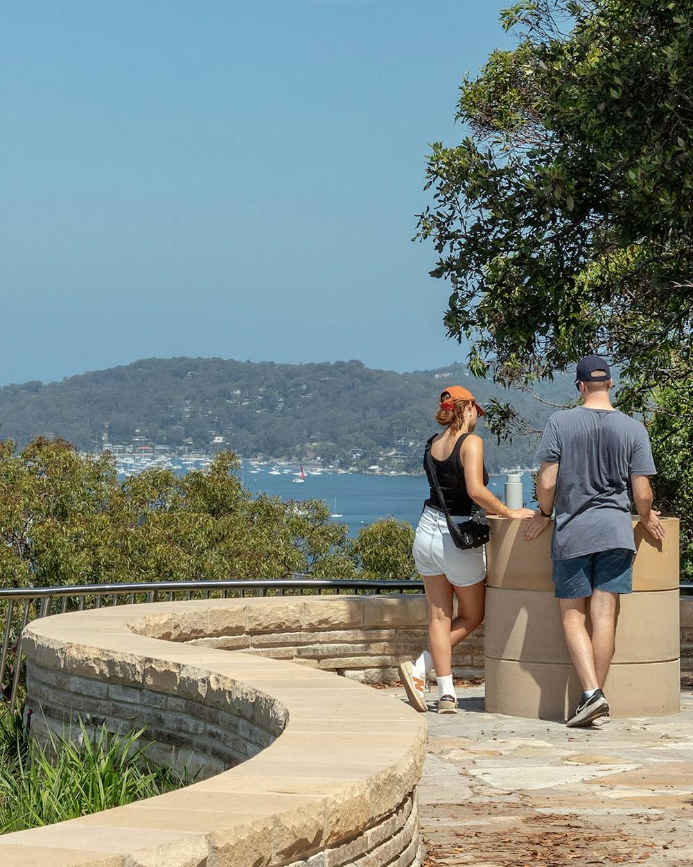 A photograph of two people at a lookout with water views on a sunny day.