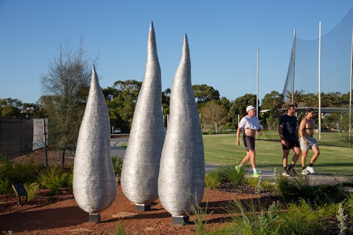 A photo of three organic, silver, sculptural forms in a park setting. Three people are walking in exercise gear on a path behind.