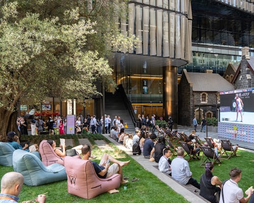 A photograph of a public, urban, green courtyard filled with people sitting and watching a tennis game on a large screen.