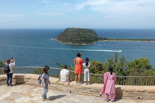 Six people taking in the water views at a sandstone lookout. Two are standing on a low wall, one is sitting on the wall, one is standing behind the wall taking a photo.