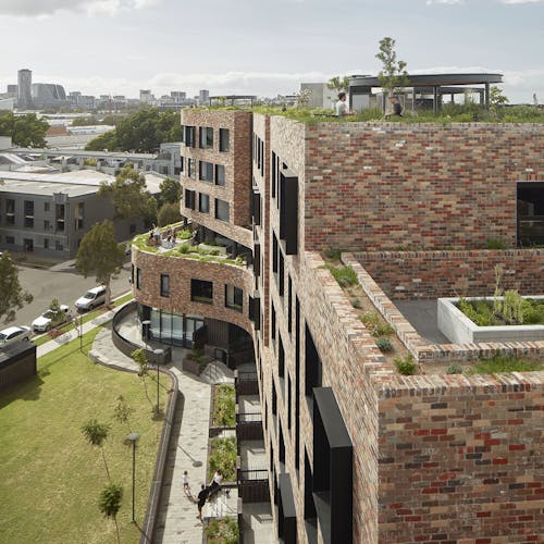 A photograph of a brick apartment building next to a pocket park, with a rooftop garden, and vegetable gardens on terraces.  
