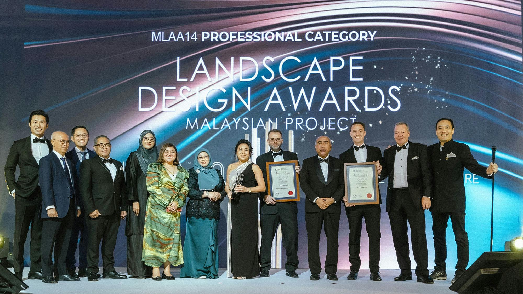 A photo of thirteen smiling people on stage receiving a "Landscape Design Award" according to the screen behind them.