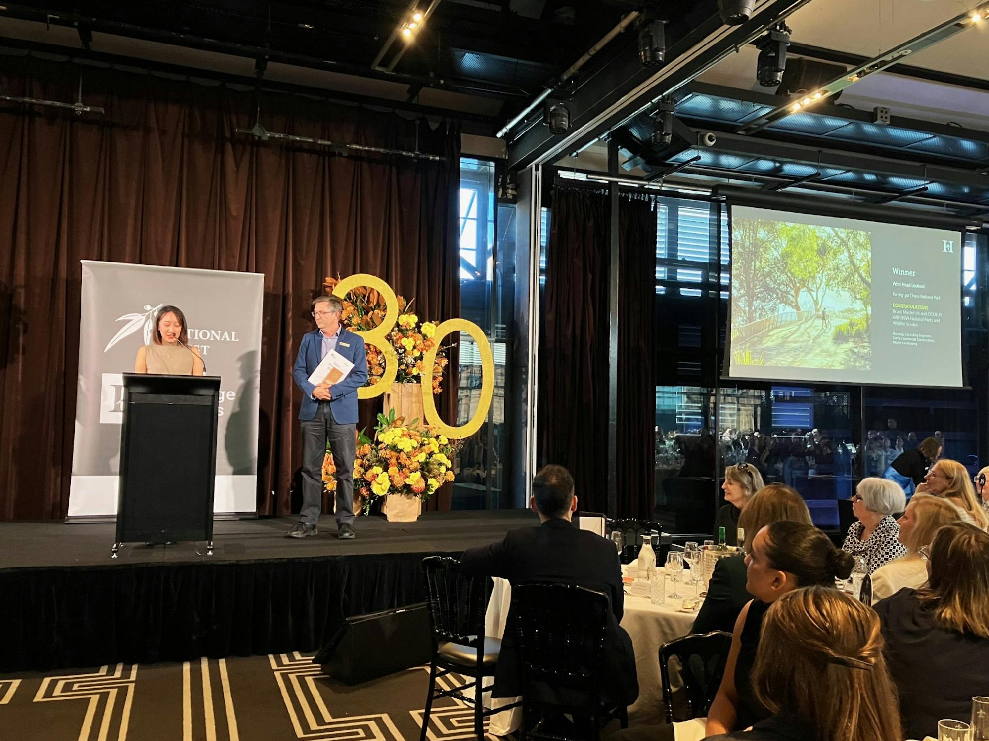 A photograph of a seated awards ceremony, two people are on stage collecting a trophy. There is a floral arrangement with a big "3" "0" cut out.