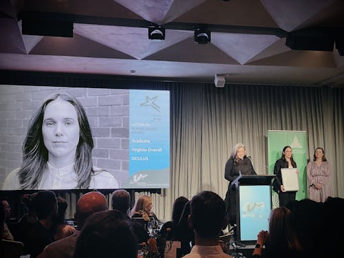A photo of an awards presentation stage. There is a black and white headshot on a projector screen, and three people on stage - one is speaking at a lectern, and one is holding a framed certificate. 