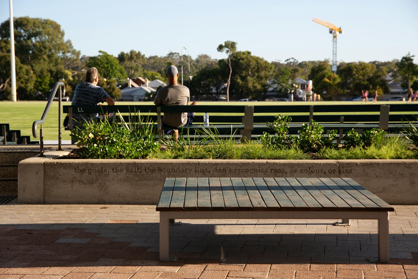 A photo of two people watching a sports field, taken from behind on a sunny day.