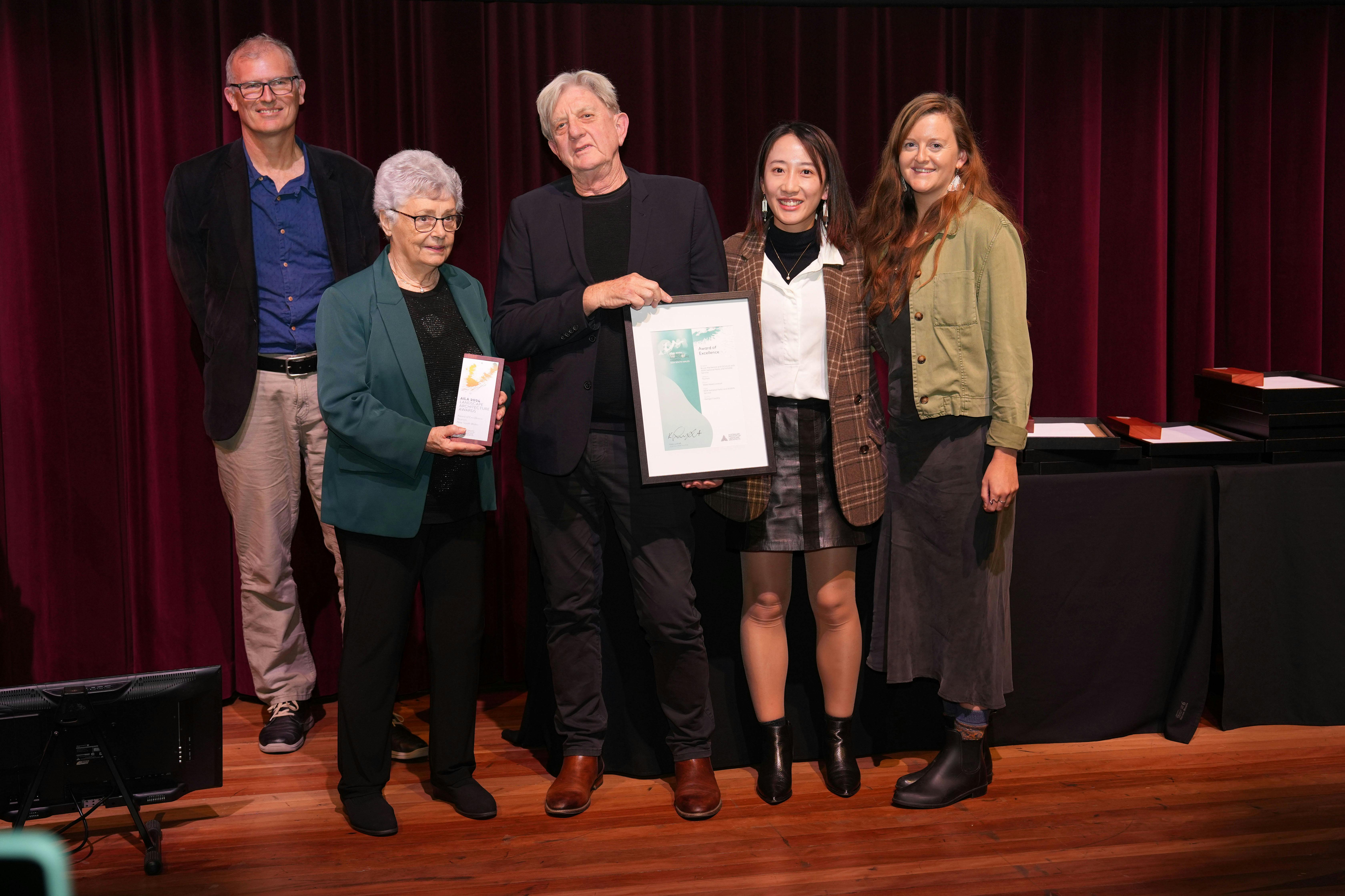 A photo of five people on stage accepting an award. One person holds a trophy, one person holds a framed certificate.