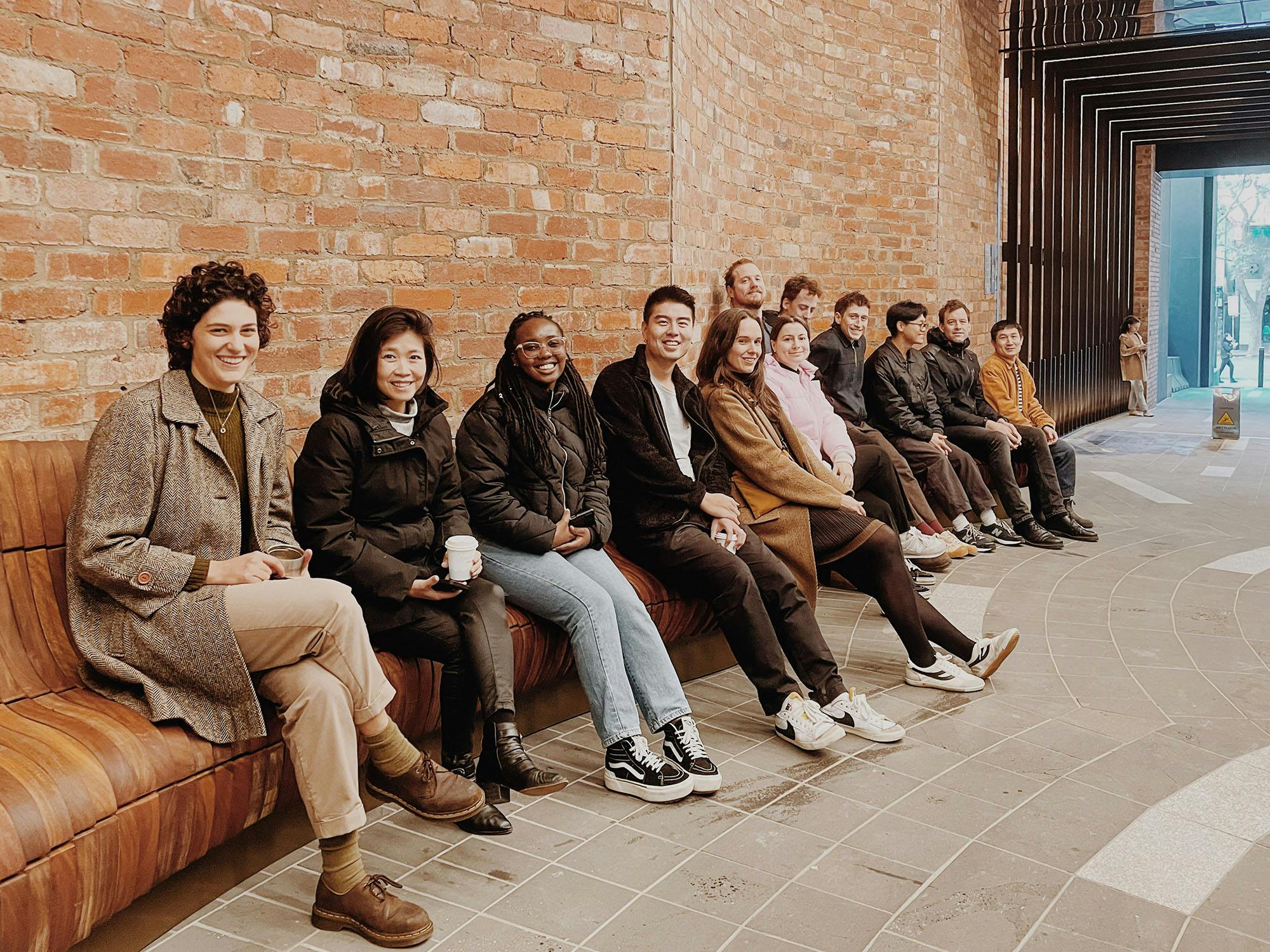 A photograph of 12 smiling people sitting on a long, curving bench.