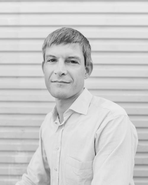 A black and white headshot of a person wearing a pale button up shirt in front of a pale roller door.