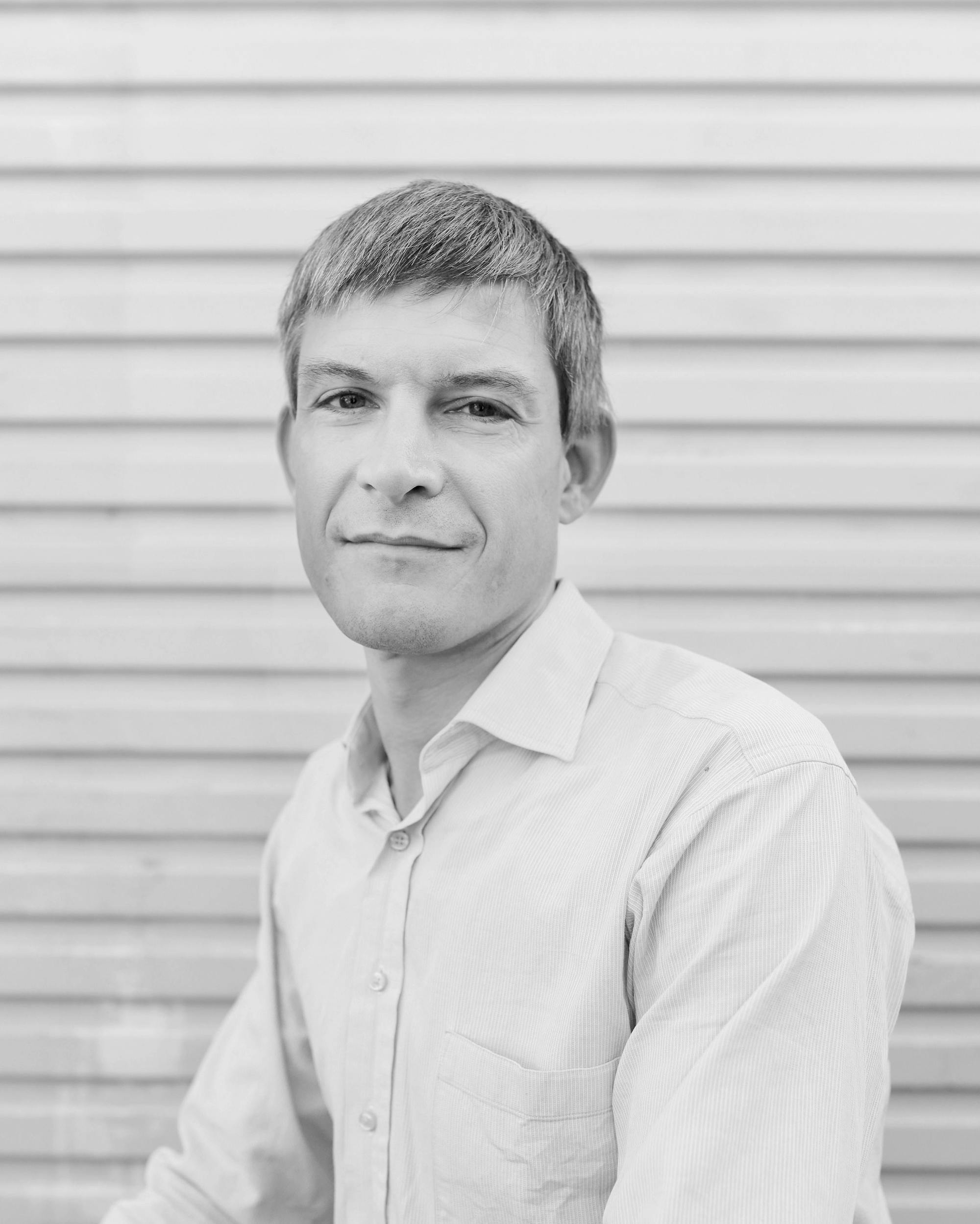 A black and white headshot of a person wearing a pale button up shirt in front of a pale roller door.