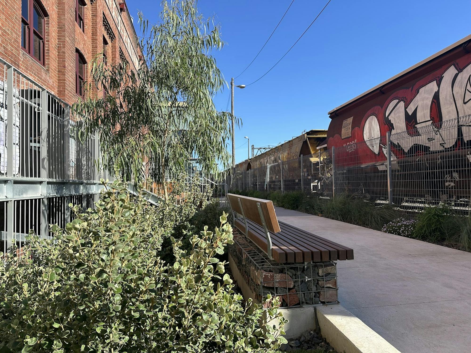 A photograph of pathways, planting, and seating between an old industrial building and railway line.