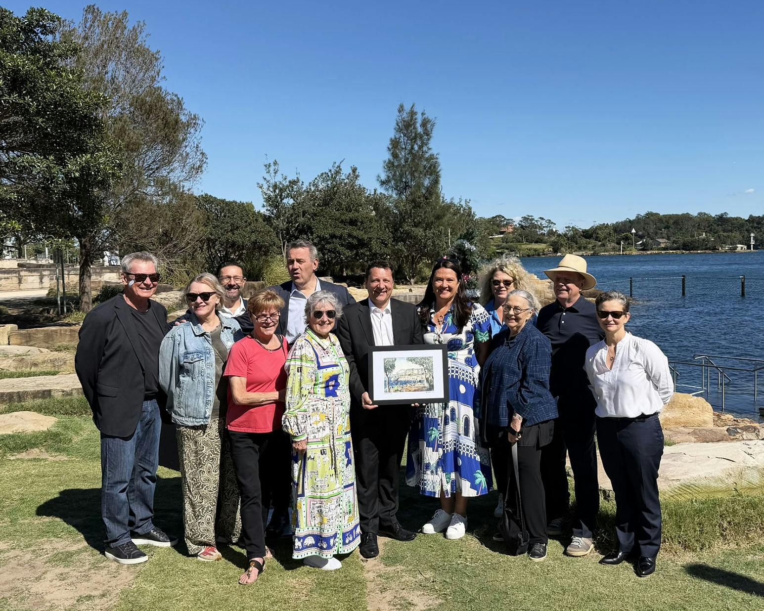 A photo of twelve smiling people posing for the camera, one in the centre holds a framed drawing of the harbour-front landscape they are standing in.