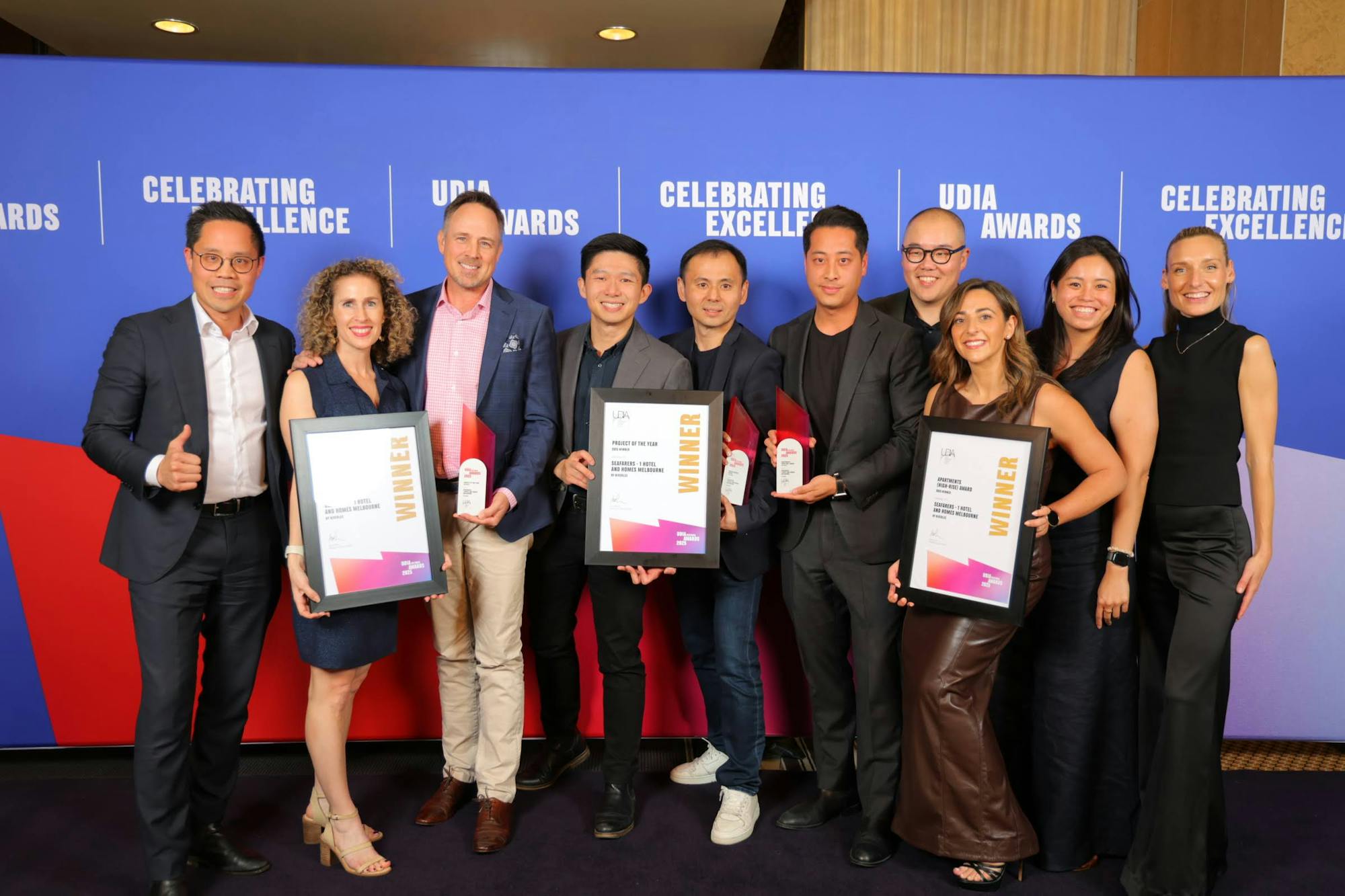 A photo of ten smiling people holding three framed certificates and three trophies. One is giving a thumbs up. 