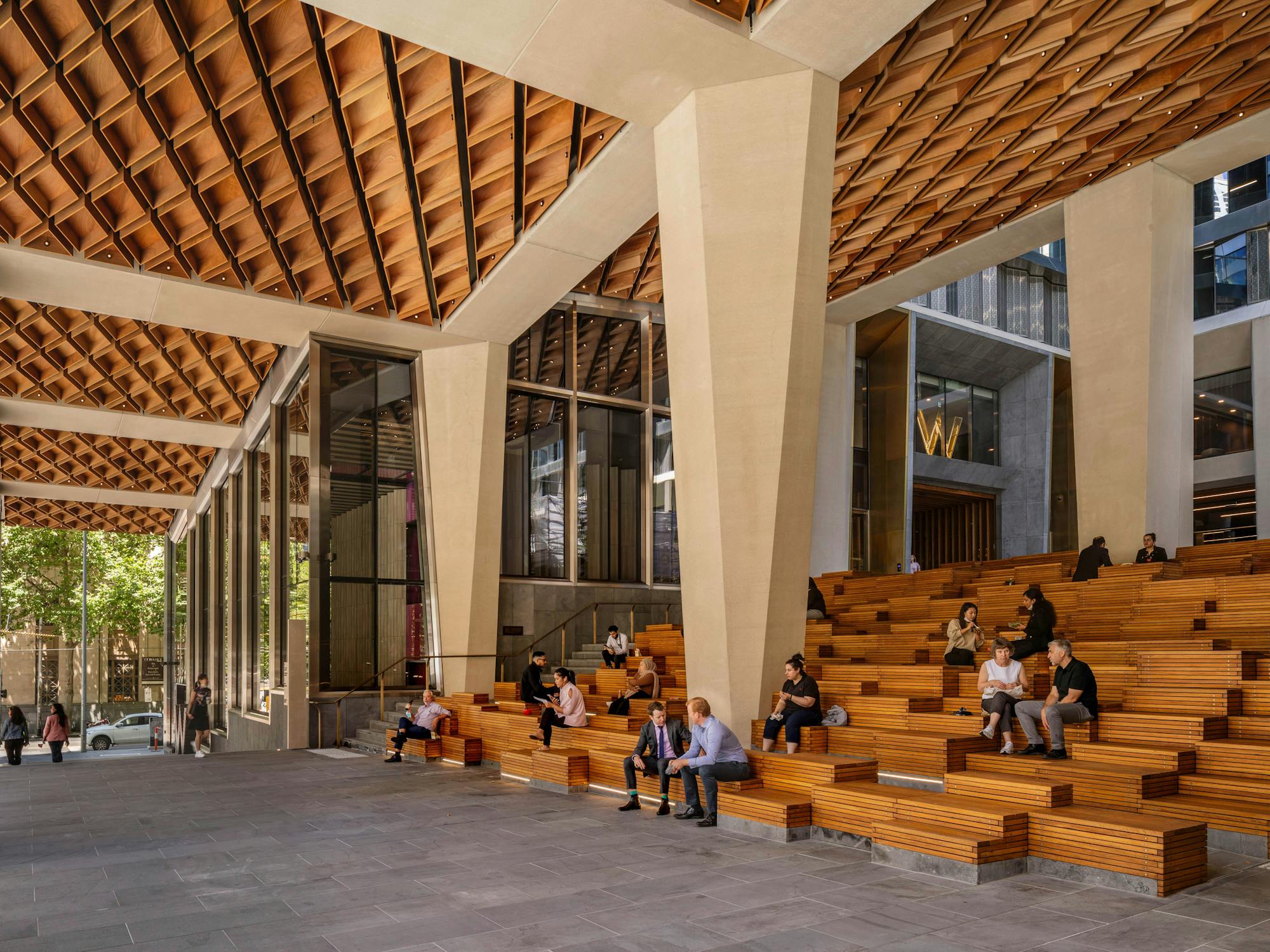 A photograph of an urban public plaza beneath a new commercial building. There are grey stone pavers and tiered timber seating. Office workers are eating lunch and talking on the seats.