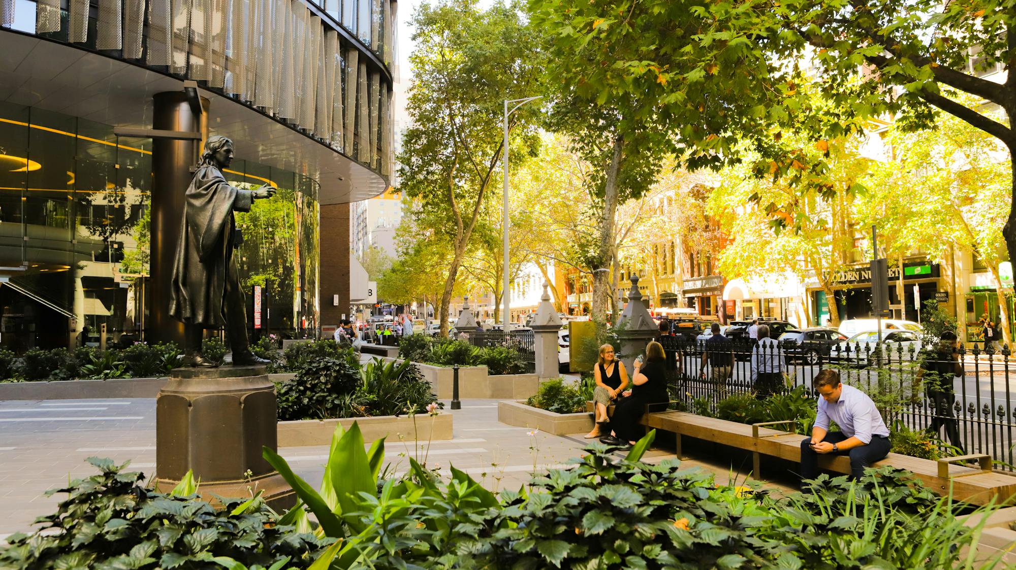A photograph of a few people sitting in an urban forecourt with planting in the foreground, tree canopy above, and a contemporary commercial building to the left. 