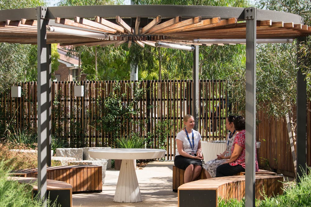 A photograph of three people in an outdoor circular seating area. There is a timber screen and shade structure, and the area is surrounded by plants and trees.