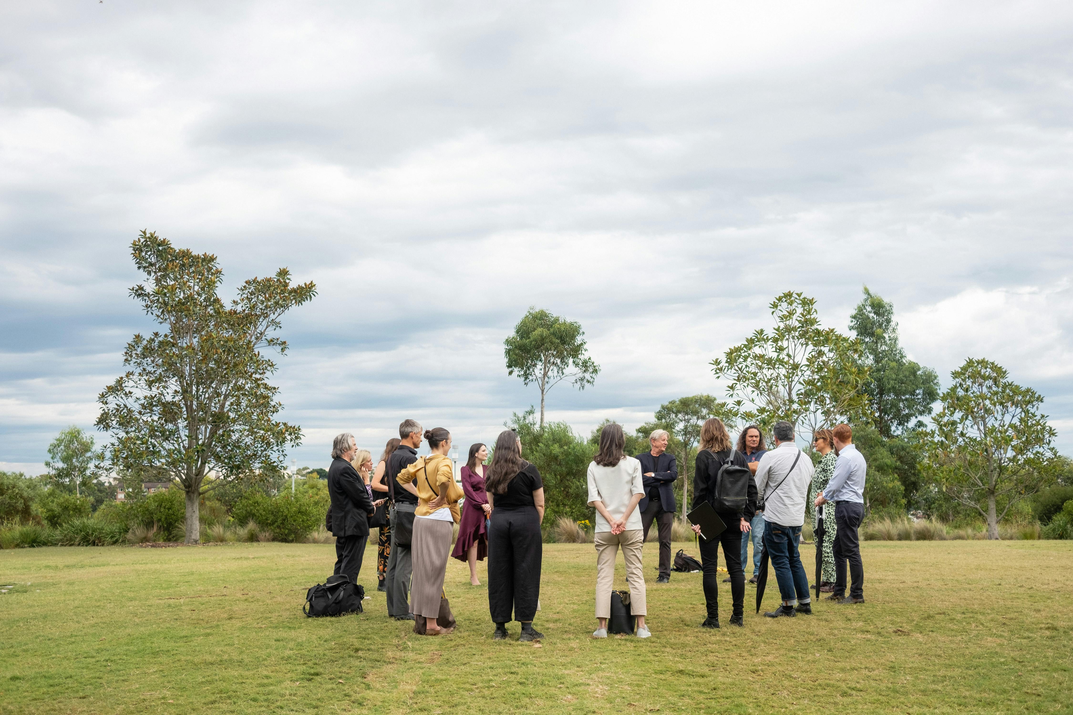 A photo of a group of people standing in a circle in a park. They are on a grass lawn, there are trees, shrubs, and an overcast sky behind them.