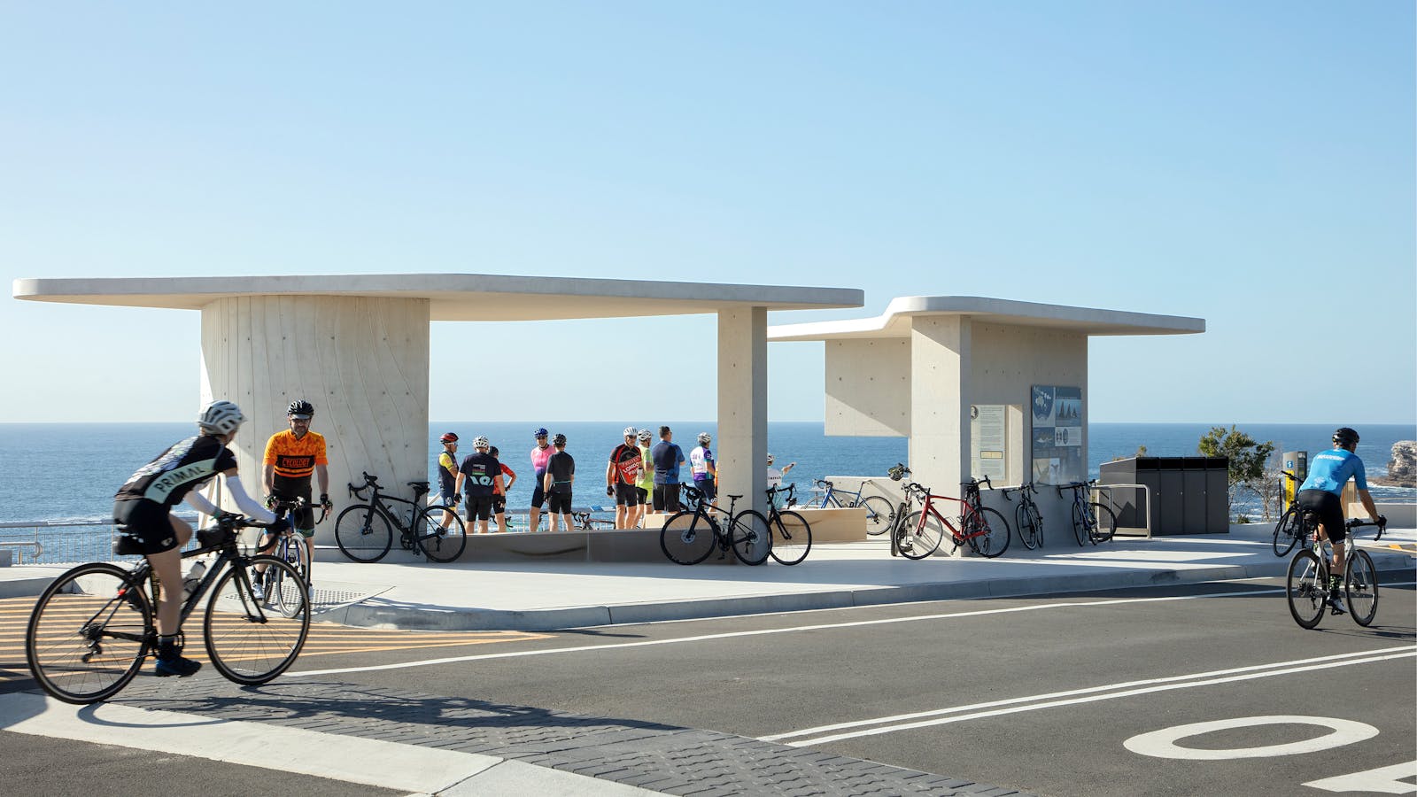 A photograph of a group of cyclist mingling around a lookout. Blue sea and Skies are behind. Two cyclists ride past on the road in front.