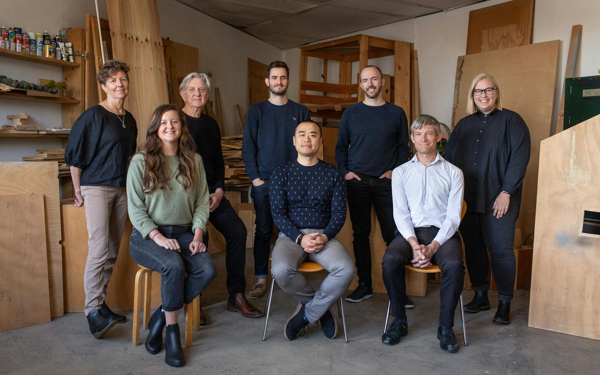 A semi-formal photograph of eight people in a studio workshop space, everyone is smilling and looking at the camera. Plywood, tools, and paint are visible in the background.