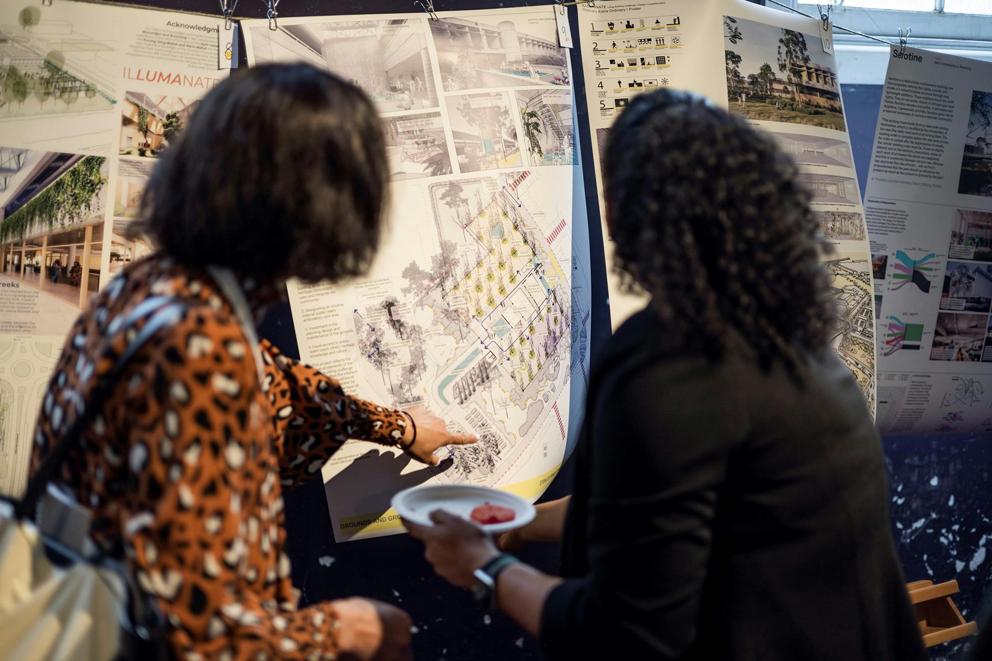 A photograph of two people from behind who are pointing at details on large posters with urban and architectural graphics.