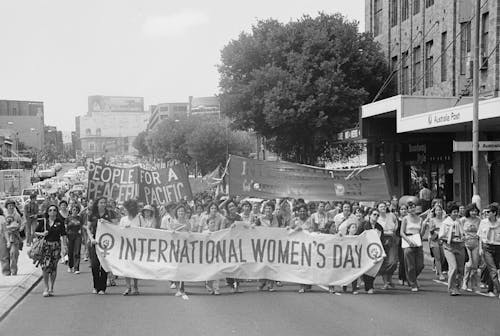 A black and white photograph (scanned from film) of an International Women's Day March in Sydney in the 1980's. People are marching towards the camera, a banner stretches across the street reading 'International Women's Day'. The crowd stretches down the street, disappearing into the background of the photo.