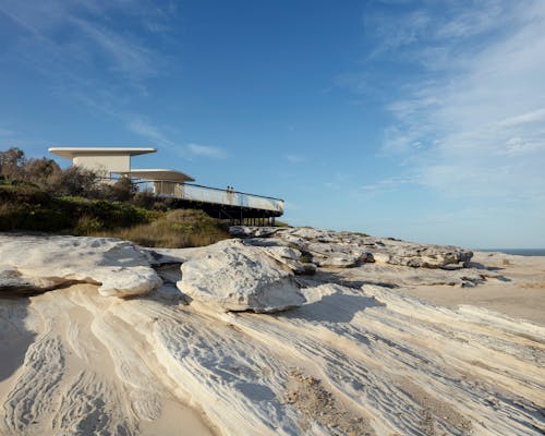 A photograph of a pale sandstone clifftop with a curved white concrete viewing shelter on top. The sky is bright blue with streaks of clouds. 