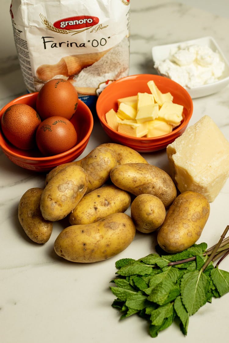 A pile of 9 potatoes on a countertop, surrounded by basil, eggs, parmesan cheese, pasta flour, and ricotta.