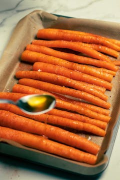 Olive oil being drizzled over halved carrots in a baking tray. 