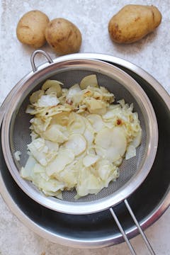 Thinly sliced potatoes and onions being strained in a sieve. 