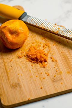on a wooden board, orange and its zest thats been grated and a greater with wooden handle