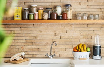 kitchen sink, on the right hand its a bucket of fresh fruit, nutribullet blender. On the left side wooden board with fresh baked bread and a bottle. Above glass jars of cooking supplies and drinking glass