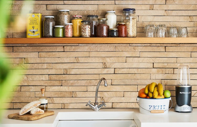 kitchen sink, on the right hand its a bucket of fresh fruit, nutribullet blender. On the left side wooden board with fresh baked bread and a bottle. Above glass jars of cooking supplies and drinking glass