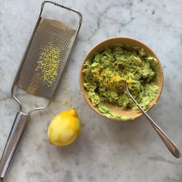 Avocado in a bowl covered in grated lemon zest. The grated lemon and the grater are next to the bowl.