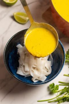 in bowl rice noodles in the bottom and yellow broth getting poured on the top 