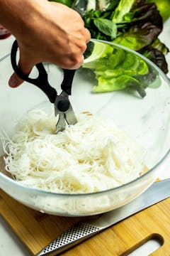 Scissors being used to cut up a bowl of vermicelli noodles, already soaked and softened.